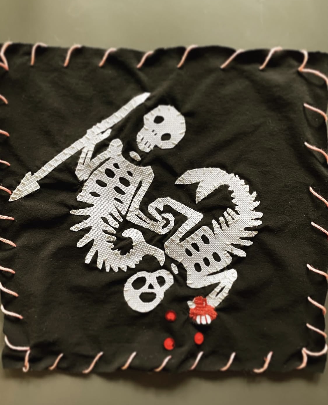 A hand embroidered flag of two mermen skeletons. One if holding a spear and the other is crushing a red heart in his boney hand. Red sequins drops squirt from the assaulted organ.