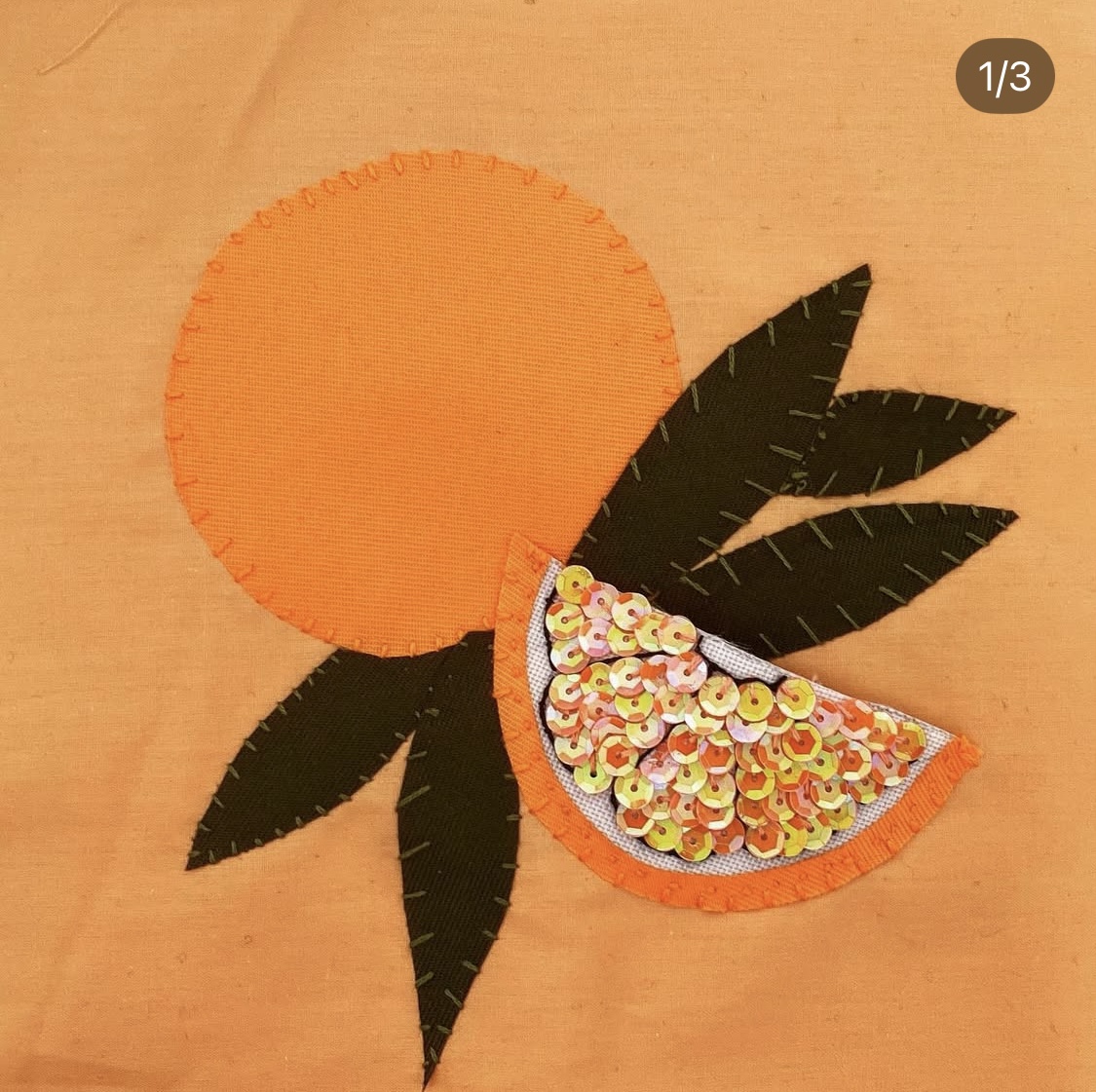 A hand embroidered flag of an orange fruit with green leaves and an orange slice, sparkling with sequins.