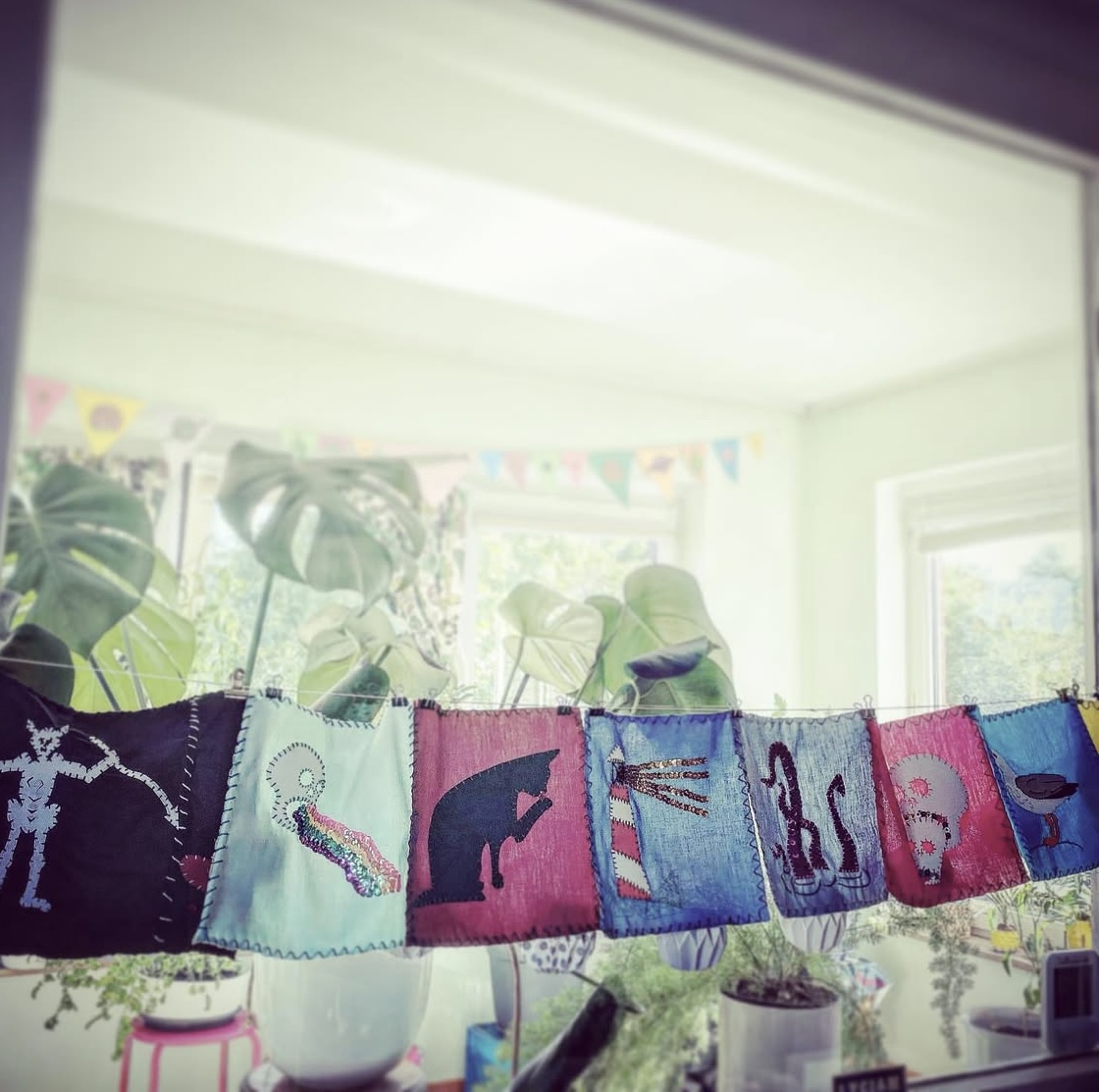 seven hand embroidered gay pirate flags are strung across a brightly lit. plant-filled window.